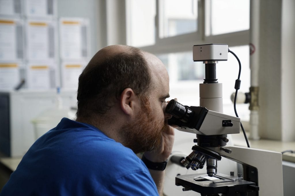 Home DE 9 Mitarbeiter der mit blauem Jansen T-Shirt vor einem digitalen Mikroskop im Labor bei der Materialprüfung für die Extrusion